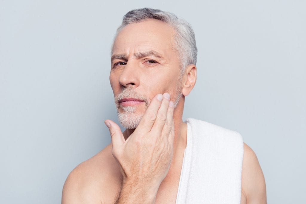 Close up potrait of a senior man analyzing his skin condition with his hand in a mirror.