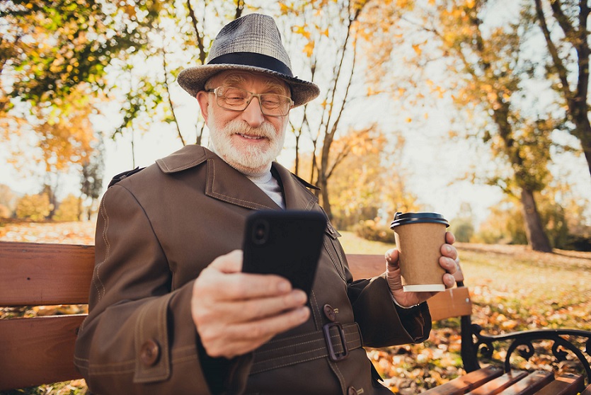 Senior man is using virtual assistance of his smartphone in park . holding a take-away coffee  cup.