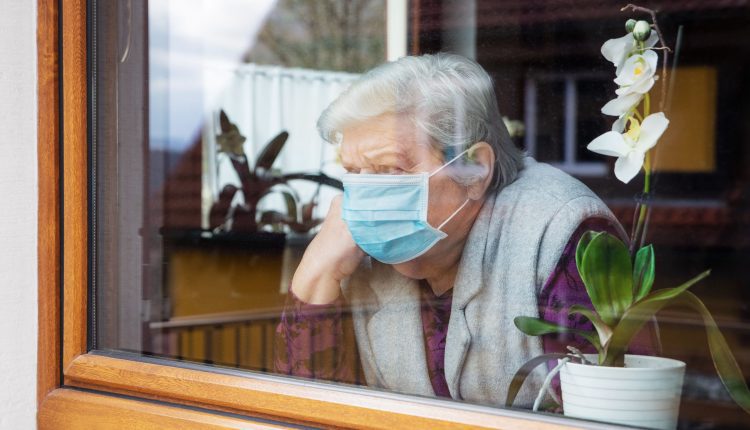 Seniors woman is in quarantine for covid-19 .and she wear blue mask and stands behind her house window