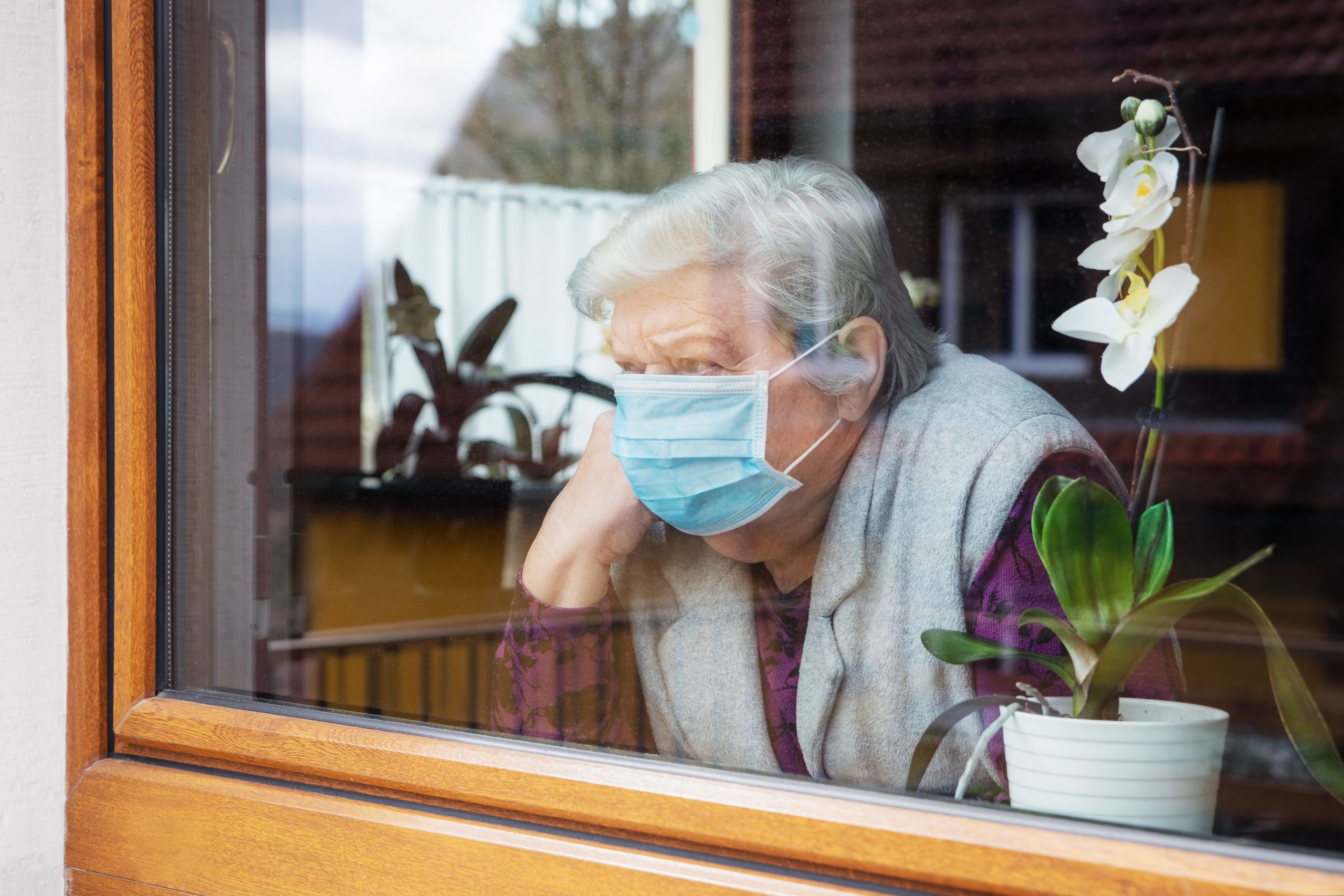 Seniors woman is in quarantine for covid-19 .and she wear blue mask and stands behind her house window