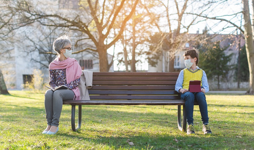 Grandmother and grandson sit separated by social distancing on park bench. The elderly avoid <a href=