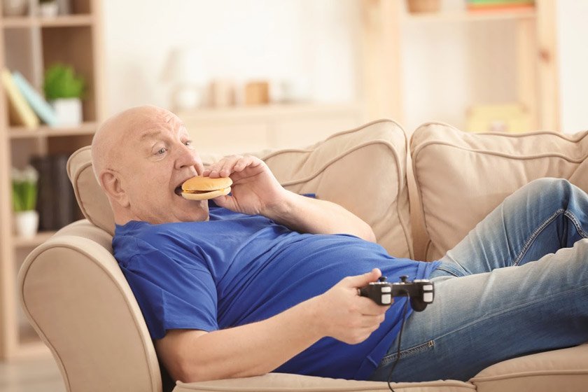 BMI for seniors: A senior man eating a burger on couch and watching T.V.  weight chart for seniors males by age and height. BMI for seneiors