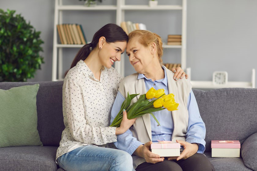 Loving young daughter hugs and gives flowers and gifts to her mature mother.