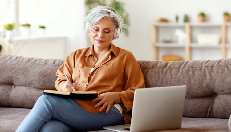 Senior woman with a headphone learning from an online course with her laptop writing down something