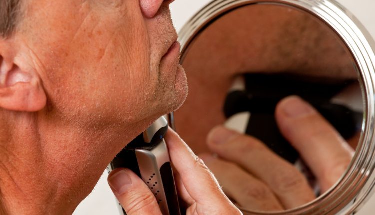 Senior man shaving his beard in front of a small round mirror