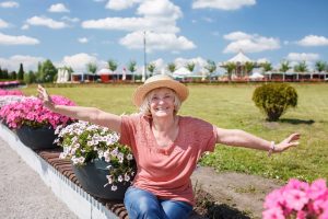 Carefree senior woman in straw hat walking in the flower park at sunny day.