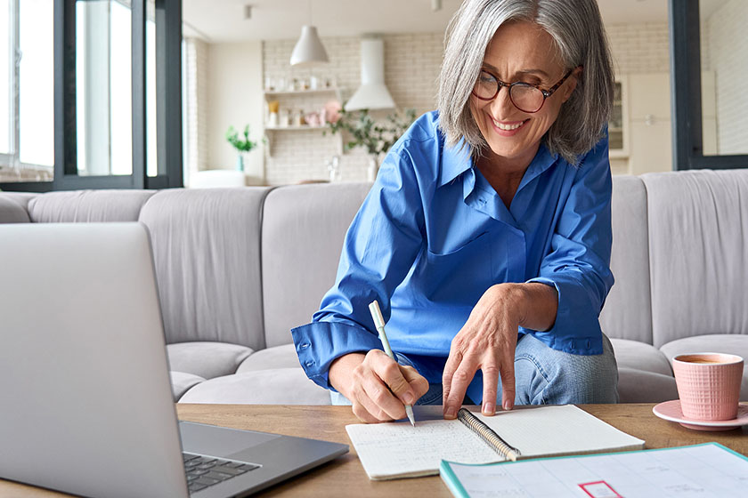 Happy mature senior woman working with her laptop from home and writing things down