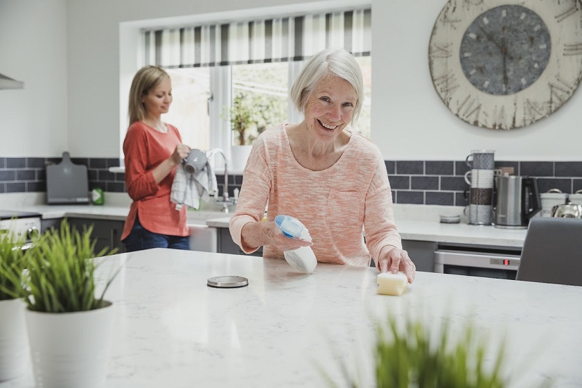 Senior woman is smiling for the camera as she cleans her kitchen with help from her daughter.