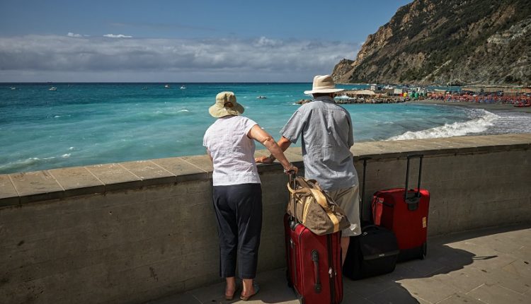 Two older adults stare at the sea with their travel bags.