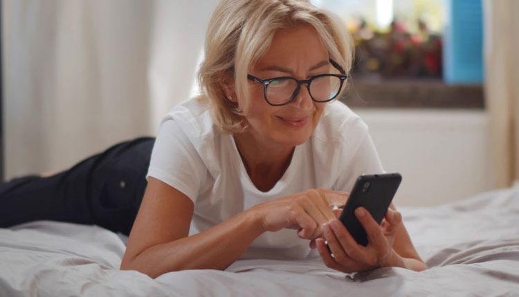 Senior woman with glasses laying on her bed and looking at her phone