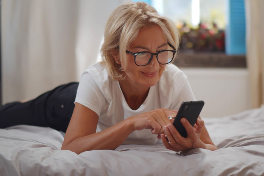 Senior woman with glasses laying on her bed and looking at her phone