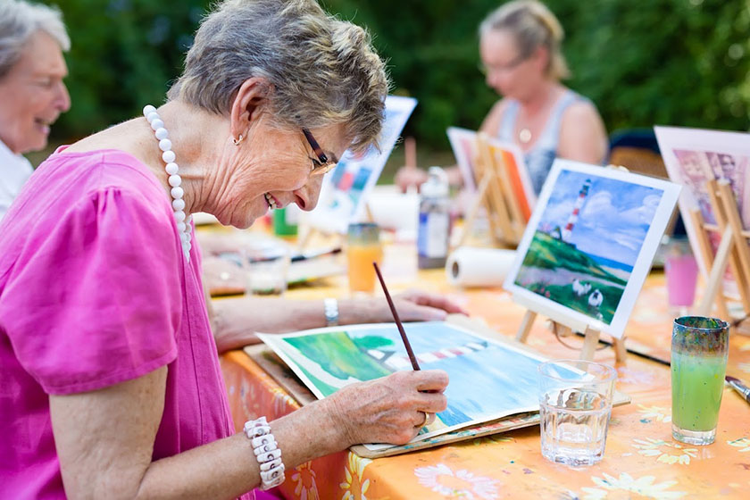 Side view of a happy senior woman smiling while doing art therapy outdoors with a group of retired women.